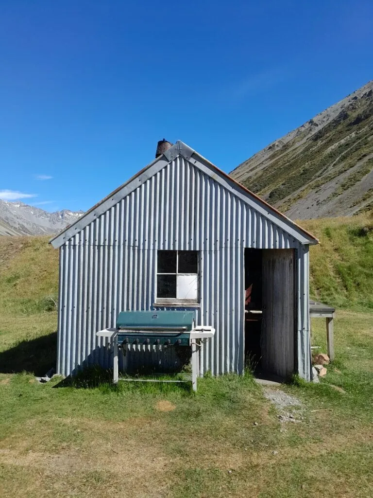 Front view of the rustic, corrugated-iron musterers' hut called Wild Mans Hut under a bright blue sky, featuring an open wooden door and a gas barbecue sitting out front in a grassy basin. Located near Top Hut in the South Branch Ashburton River, Hakatere Conservation Park, Canterbury