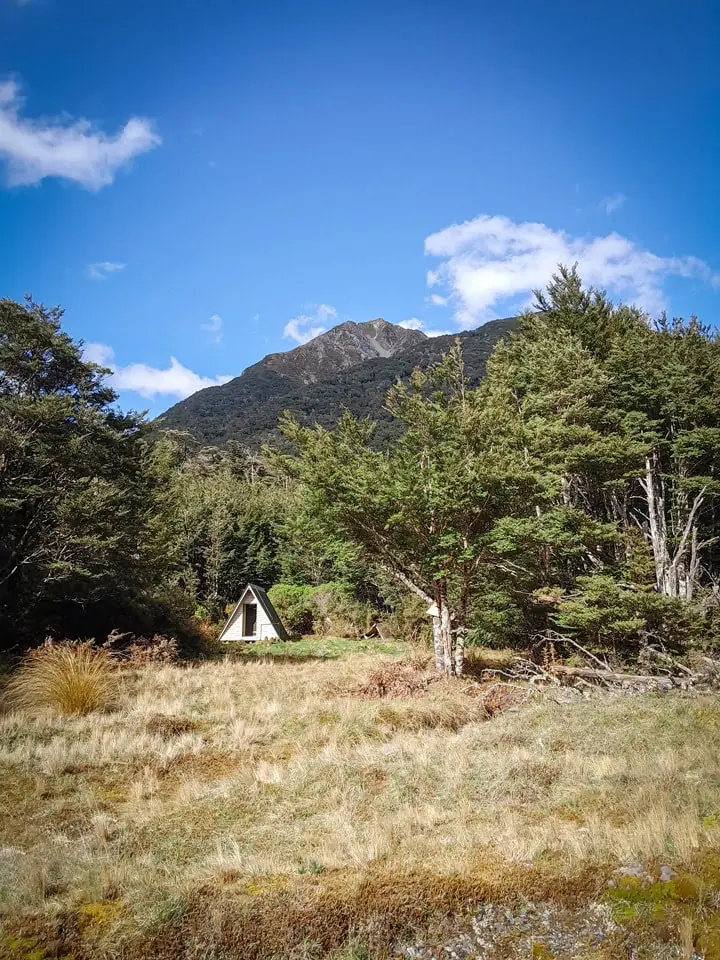 A small, silver A-frame Worsley Biv sits in a tussock clearing surrounded by a dense beech forest with a prominent rocky mountain peak rising behind it in the Poulter Valley, Arthur's Pass.