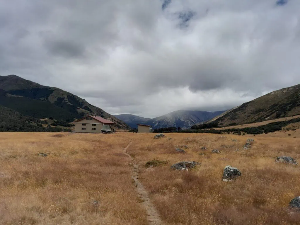 Large backcountry hut, Anne Hut, in an open golden tussock valley on the St James Walkway under a moody, overcast sky, St James Conservation Area.