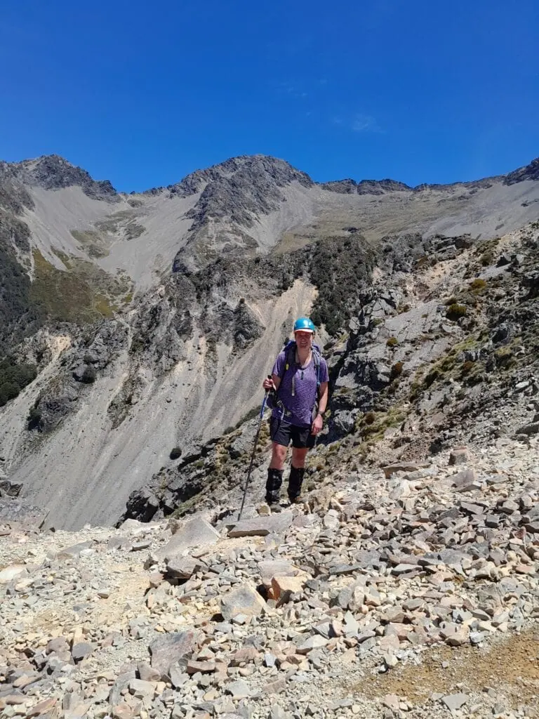 Hiker with a helmet, backpack, and trekking poles standing on a loose scree slope with the summit of Bounds Peak in the background, Marlborough