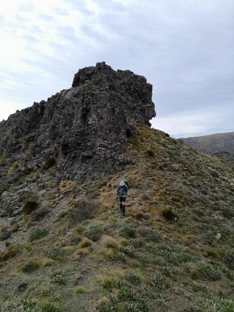 Hiker navigating around a large rocky outcrop on the steep trail ascending toward Blairich peak above Black Birch Bivvy in Ferny Gair Conservation Area, Marlborough
