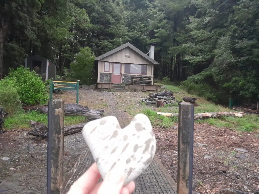 Hand holding a heart-shaped stone in front of Coldwater Hut on the edge of Lake Rotoiti in Nelson Lakes National Park.