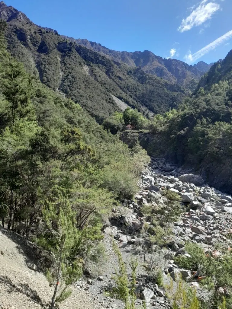 Distant view of pink Gosling Hut perched on a high river terrace surrounded by native bush and rugged mountains.