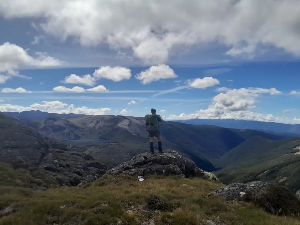 Tramper with a backpack standing on a rocky outcrop enjoying wide views of mountain ranges and valleys along the Lockett Lakes Loop including Lake Lockett