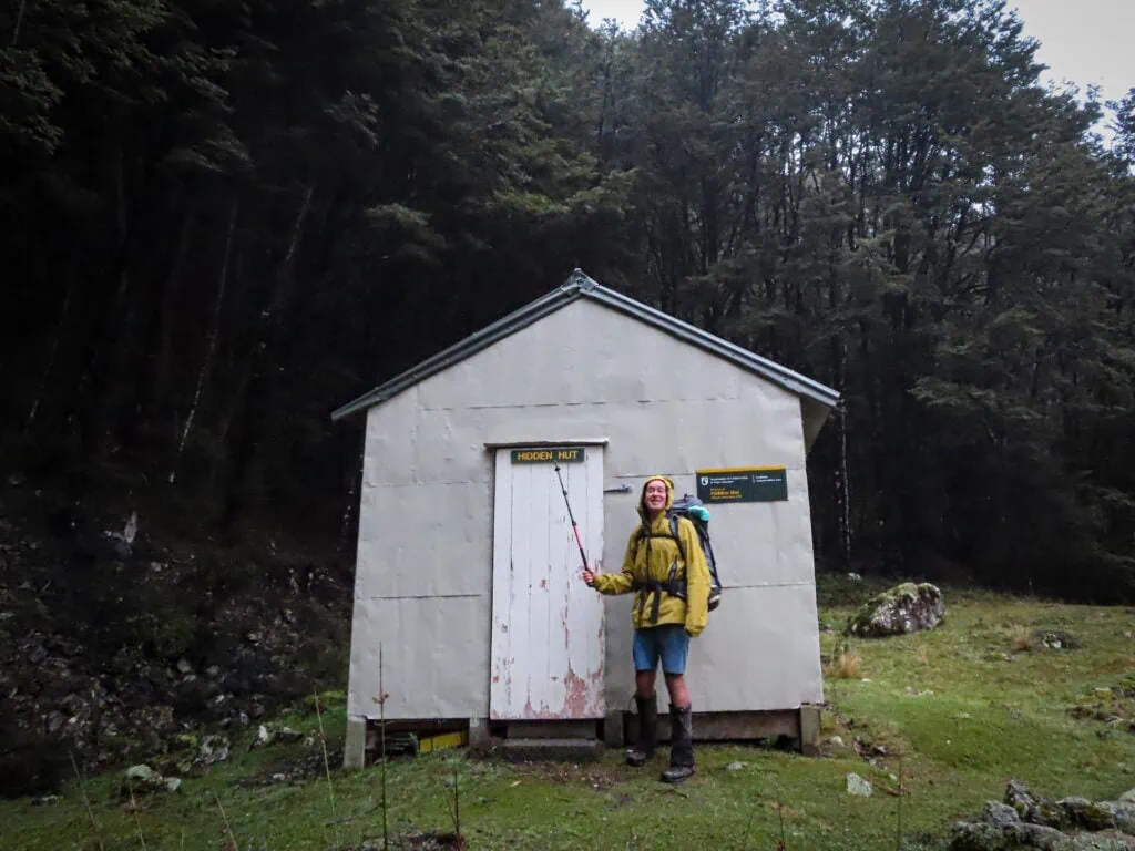 Hiker wearing a yellow coat, standing outside a white hut, pointing at the name Hidden Hut with his walking pole