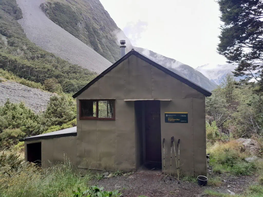 Front view of Hopeless Hut surrounded by native forest and alpine mountains in Nelson Lakes National Park.
