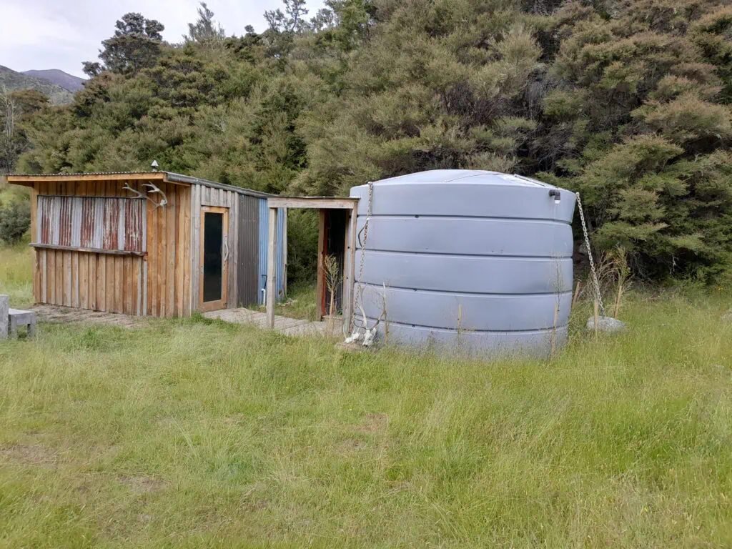 Wooden Stronvar Station Hunting Hut with corrugated iron siding and a large plastic water tank set in a grassy clearing on the route to Gosling Hut