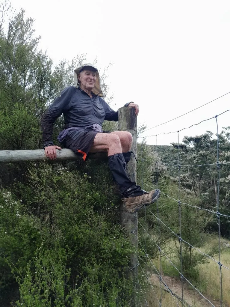Tramper wearing gaiters climbing over a high wire deer fence while traversing through Ferny Gair Conservation Area, Awatere Valley, Marlborough