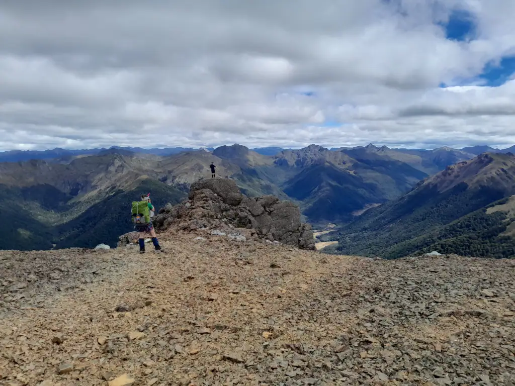 Tramper standing on a rocky outcrop on Iron Hill enjoying wide views of mountain ranges and view down into the Cobb Valley