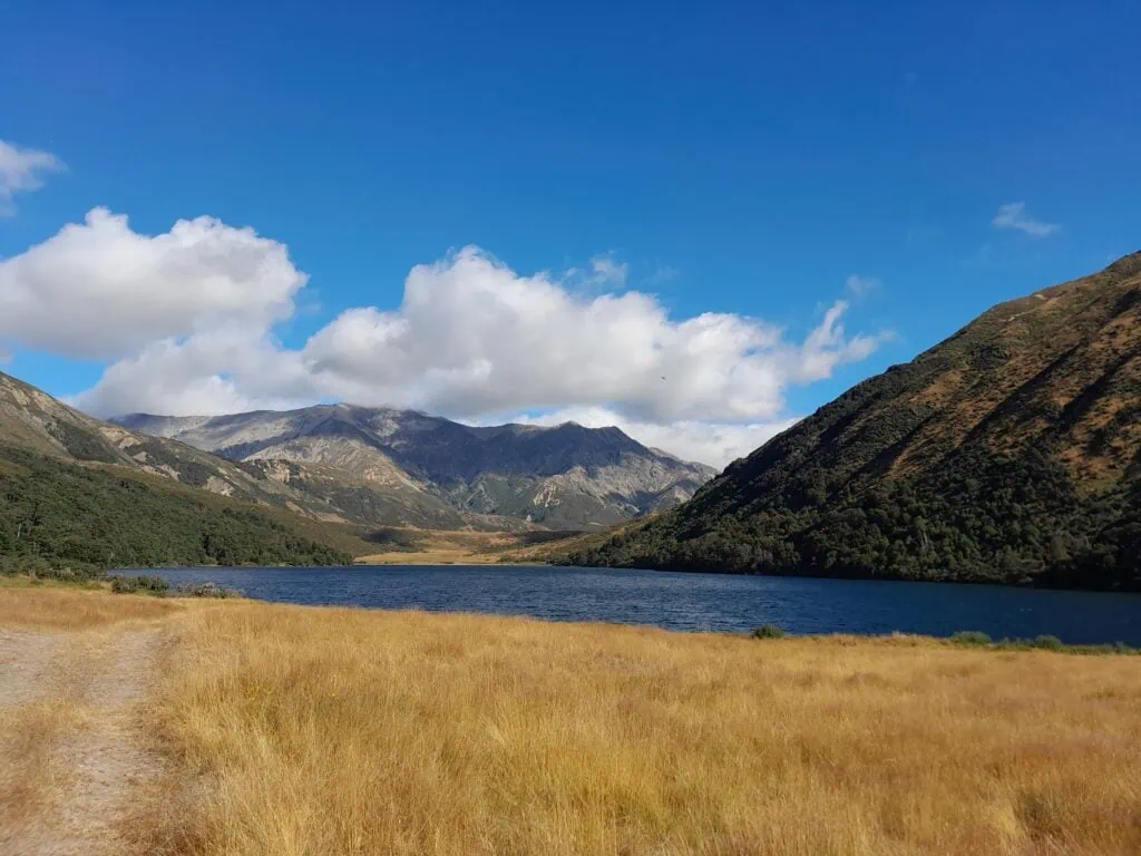 Deep blue waters of Lake Guyon bordered by golden tussock grass and rolling mountain hills under a partly cloudy sky, St James Conservation Area