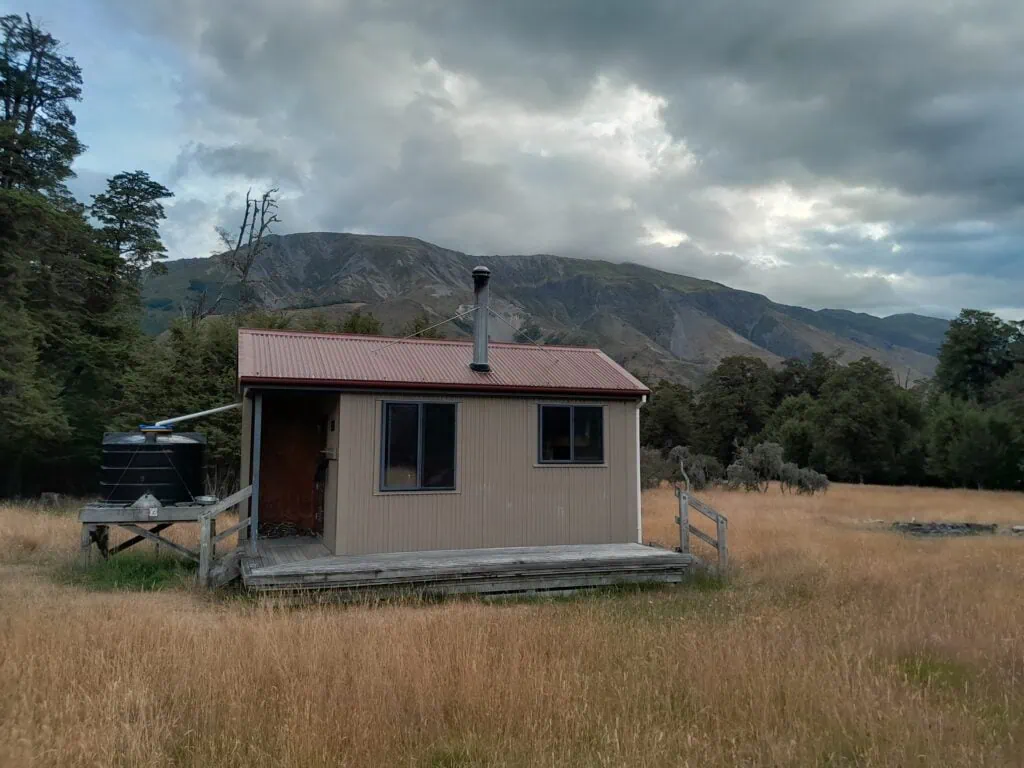 Magdalen Hut, a modern wooden backcountry hut with a rainwater tank, situated in a grassy field under dramatic clouds on the St James Walkway, St James Conservation Area