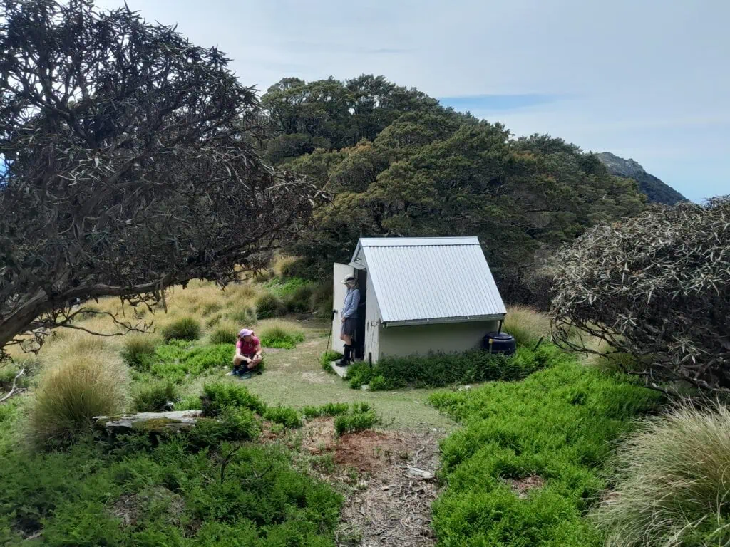 Hikers resting outside Mt Sunday Bivvy shelter surrounded by subalpine scrub and native bush, Richmond Forest Park, Marlborough