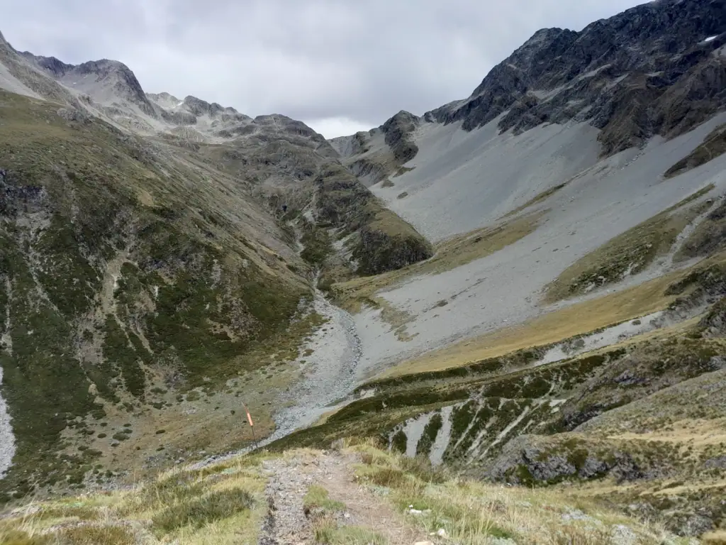 Hiking trail descending toward a rocky riverbed framed by steep alpine scree slopes and rugged mountain peaks with Lake Thompson hidden from view on the Waiau Pass Route, St James Conservation Area & Nelson Lakes National Park
