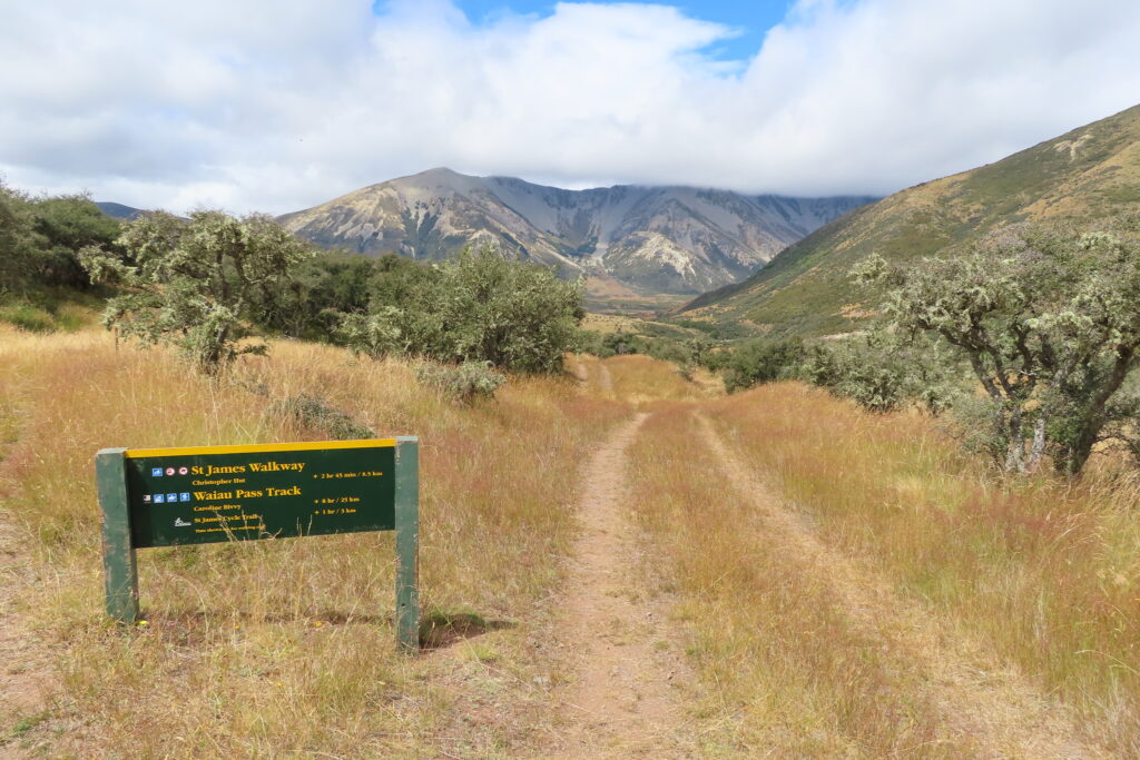 Green Department of Conservation directional sign marking the St James Walkway and Waiau Pass Track along a dirt 4WD road, Nelson Lakes National Park