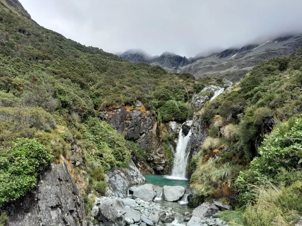 Cascading mountain waterfall flowing into a clear turquoise pool surrounded by dense alpine scrub and misty peaks on the climb on the Waiau Pass Route, St James Conservation Area & Nelson Lakes National Park.