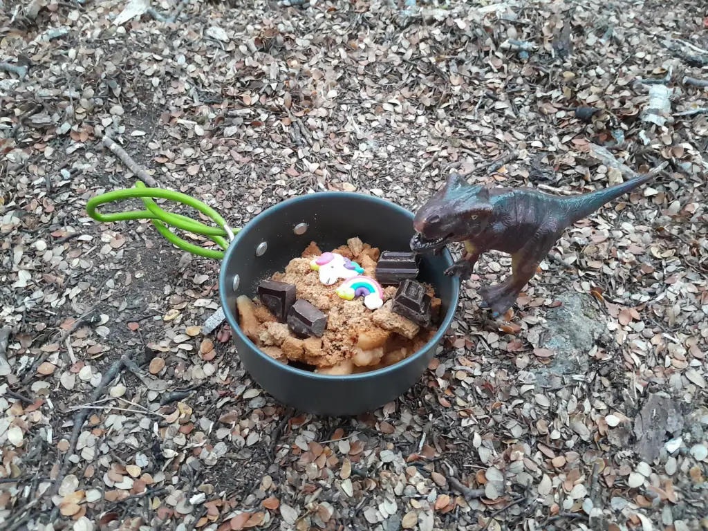 Deconstructed apple crumble in a camping pot topped with chocolate, unicorn sprinkles, and a toy dinosaur at Lake Lockett
